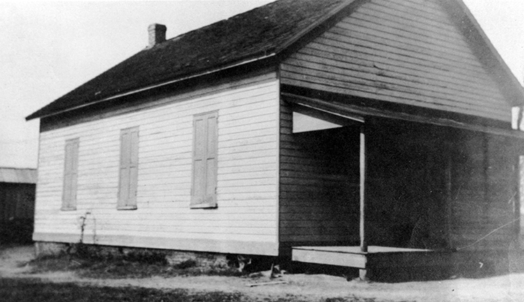 Black and white photograph of the Groveton School. The building is a small, one-story structure with a brick chimney in the rear. There is a porch and awning over the front entrance. The exterior siding has been painted white. There are three windows visible on one side, with wood shutters that are closed.