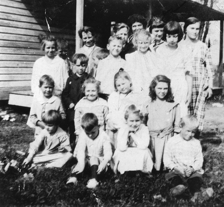 Black and white photograph of 18 children and their teacher posed in front of the Groveton School. The children are of various ages, some as young as age six and others in their teens.