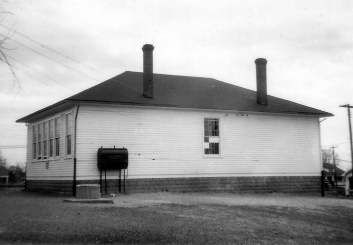 Black and white photograph of the two-room Groveton School. The rear of the building is shown. The school was heated by oil, and the oil reservoir can be seen on the side of the building. A cement well is located nearby. 