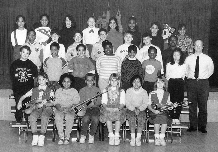 Black and white photograph of Groveton Elementary School's Strings musical performance group from the 1989 to 1990 yearbook. 28 children and one adult are pictured. The children are arranged in four rows on risers in front of the school's stage. The children in the first row are seated and are holding violins.