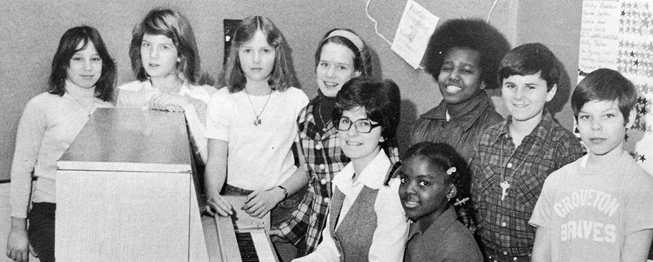 Black and white photograph of Groveton Elementary School's Choral Festival Group from the 1976 to 1977 yearbook. Eight children and one adult are pictured. The adult and one student are seated at an upright piano. The remaining students surround them.