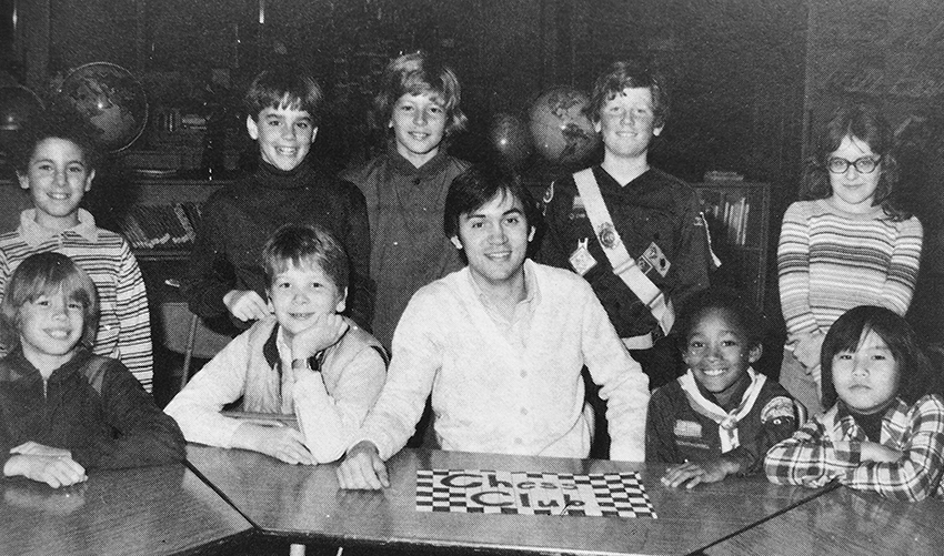 Black and white photograph of Groveton Elementary School's Chess Club from the 1980 to 1981 yearbook. Nine children and one adult are pictured. Four of the children and the adult are sitting at a table in what appears to be the library. There is a sign on the table in front of them that reads Chess Club. The sign has been decorated to look like a chess board.