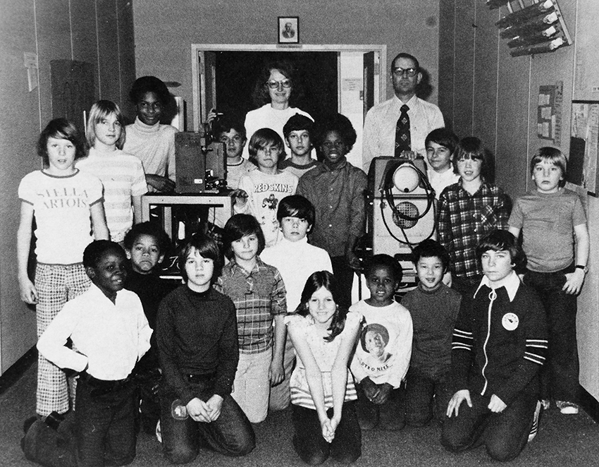 Black and white photograph of Groveton Elementary School's Audio-Visual Committee from the 1981 to 1982 yearbook. 19 children and two adults are pictured. They are posed next to a film projector on a cart and another, unidentified, piece of electronic equipment on a second cart. Some of the children are standing, others are kneeling on the floor. The two adults are visible in the far back.
