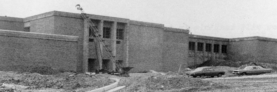 Black and white photograph from Groveton Elementary School's 1971 to 1972 yearbook showing construction progress on the new Groveton School. The original main entrance of the building is shown. The building is a two-story brick structure. Construction equipment is visible near the school's entrance, and two cars are parked in front of the building. The grounds have yet to be landscaped and there are mounds of dirt visible in the foreground.