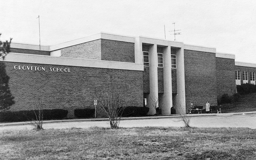 Black and white photograph of Groveton Elementary School taken in 1983. The original main entrance to the building is shown. Two adults, possibly senior citizens, are seated on benches near the main entrance.