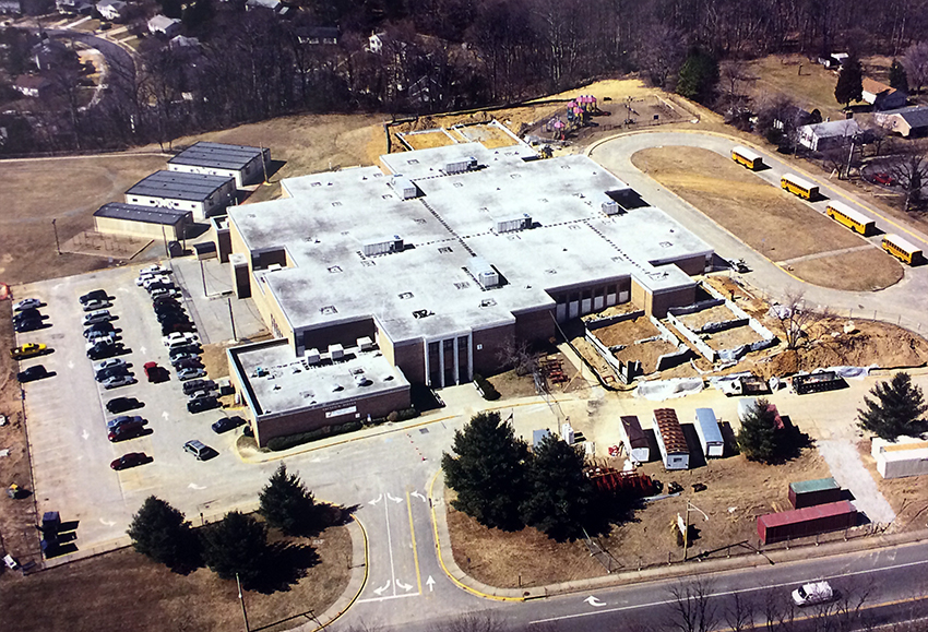 Aerial photograph of Groveton Elementary School, taken from above Harrison Lane looking toward the school from the east. Several trailers are visible behind the building. The foundations have been laid and cinderblock walls are going up on additional classroom wings on the east and west sides of the building. A portion of the driveway near the old main entrance is blocked off and construction trailers and equipment are staged in this area.