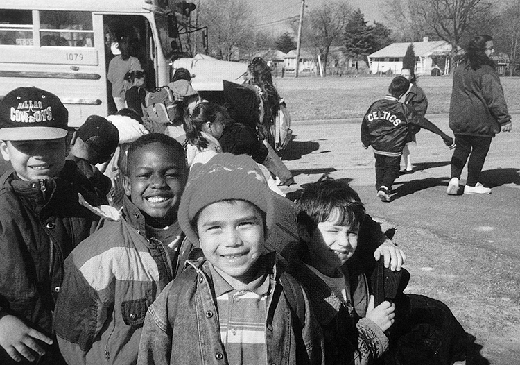 Black and white photograph of students waiting to board a school bus during the 1993 to 1994 school year. Four boys at the back of the line have turned around and are smiling broadly for the camera. They are wearing coats and hats. There are no leaves on the trees in the background, and several houses are visible in the far distance.