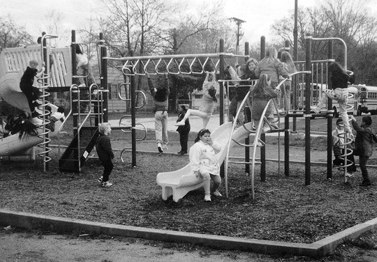 Black and white photograph of the new playground equipment. There is a spiral-shaped slide, spiral ladder, monkey bars, a curved slide, a curved ladder, and two raised platforms. 16 children are visible playing on the equipment. Two school buses are parked in the far distance.