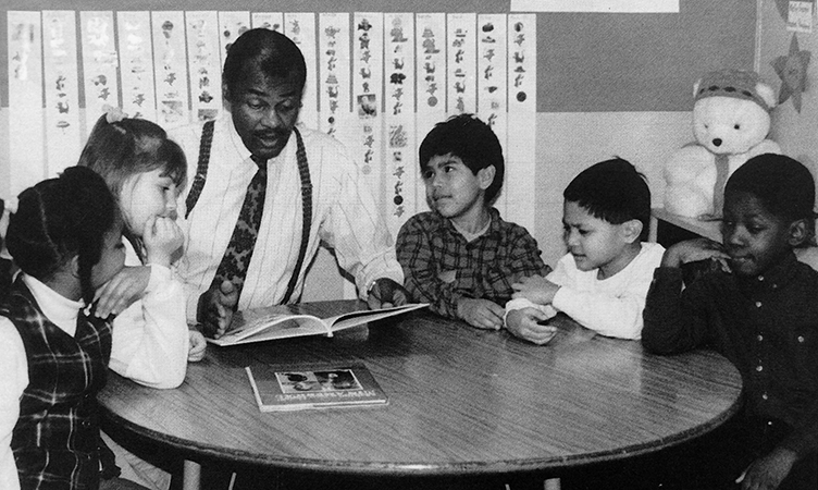 Black and white photograph Principal Thompson reading to a group of students. Five children, three boys and two girls, are seated with Thompson at a small round table in a classroom.