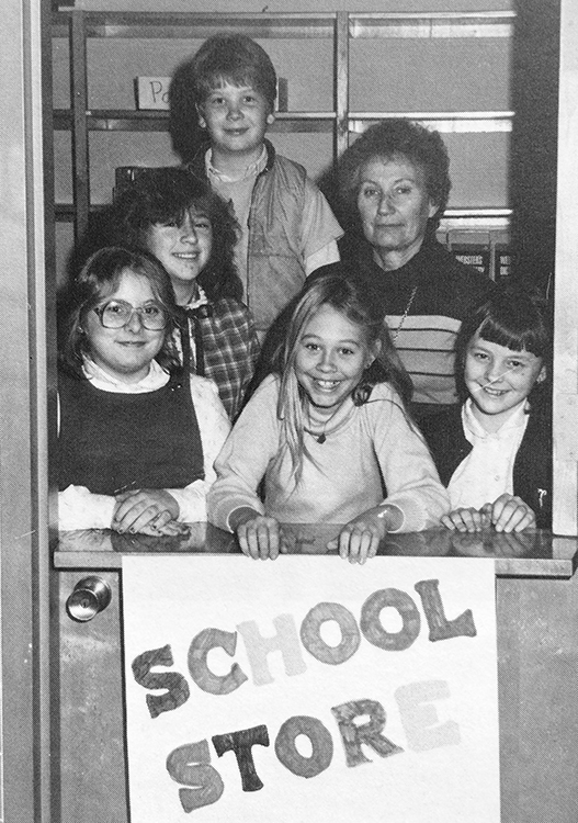 Black and white photograph of Groveton's School Store Committee from the 1980 to 1981 yearbook. Five children and one adult are pictured. They are standing inside the school store. The door to the store is a Dutch door, with a top half that opens separately. The top half is open and the bottom is closed. A girl in the center is holding a sign in front of the door that reads School Store.