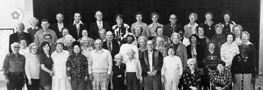 Black and white photograph of some of the senior citizens who took part in the nutrition program during the 1976 to 1977 school year. 48 people are in the picture. They are standing in four rows in front of the school stage. Two ladies are seated in chairs in the foreground.