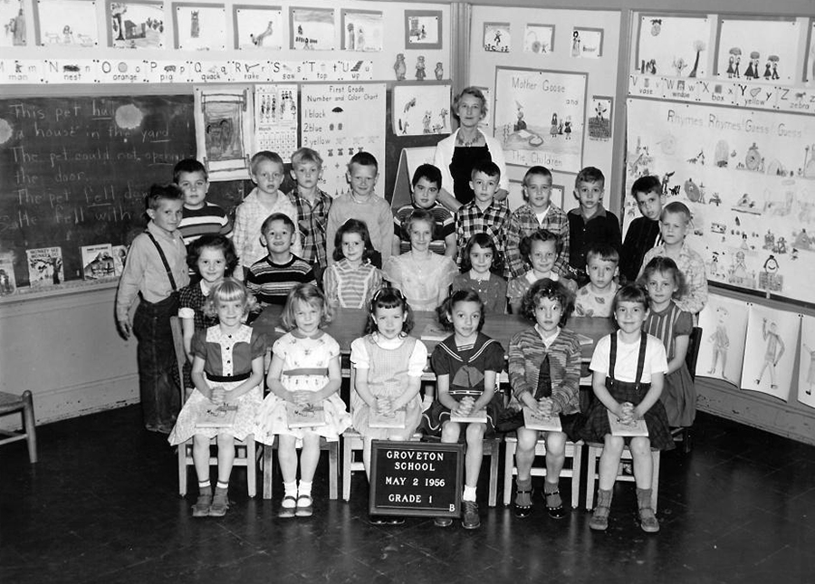 Black and white photograph showing a first grade class portrait taken on May 2, 1956. 25 children and their teacher are shown. The children are arranged in three rows in the corner of their classroom. The teacher is standing at the back of the group. The walls are heavily decorated with student artwork. The chalkboard is visible on the far left.