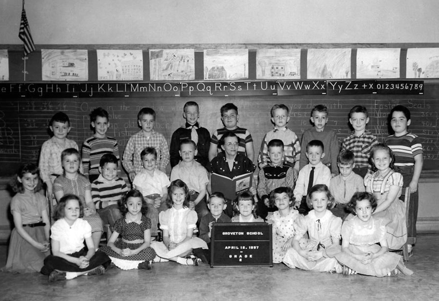 Black and white photograph of Mrs. Hare’s second grade class taken on April 16, 1957. 26 children and their teacher are shown. The children are arranged in three rows in their classroom in front of the chalkboard. Mrs. Hare is seated in the middle of the group holding a book in her lap.