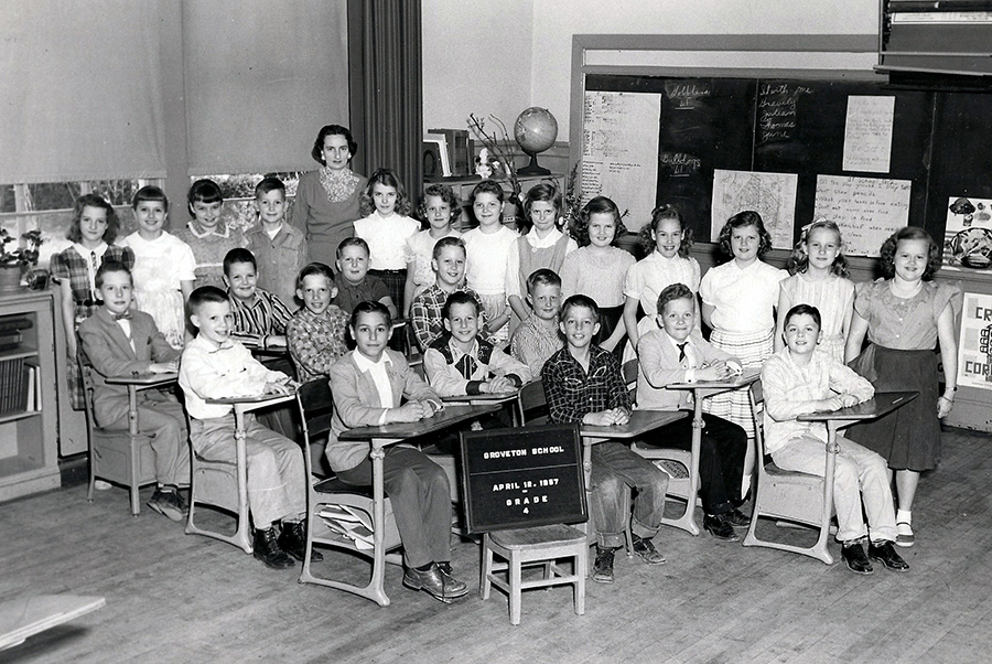 Black and white photograph of Mrs. Higginbottom’s fourth grade class taken on April 16, 1957. 25 children and their teacher are pictured. The boys are all seated at desks that have been pushed to one corner of the room. The girls and the teacher are standing behind the desks. A globe and chalkboard are visible behind the children.