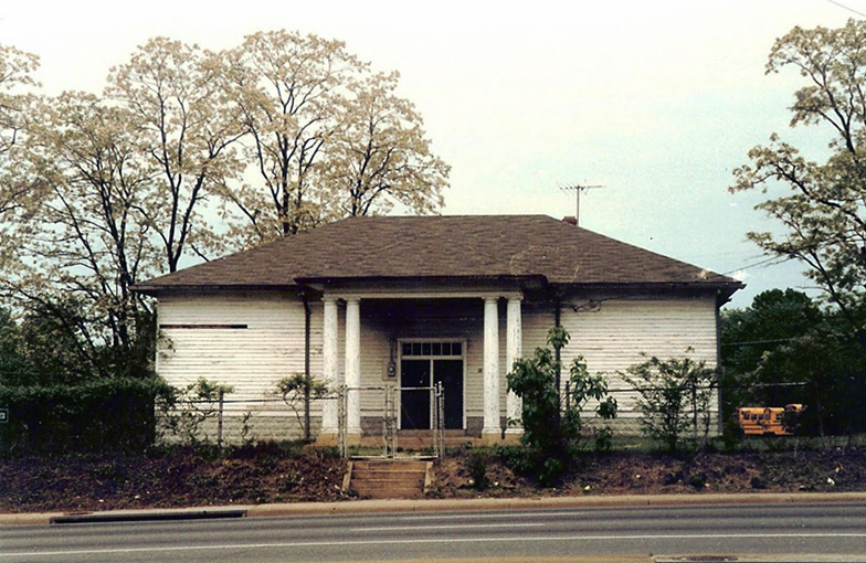 Color photograph of the two-room Groveton School taken prior to demolition. The picture was taken from across Route 1 looking toward the front of the building. A small set of stairs leads up from the sidewalk along Route 1 toward the building. The school yard is fenced and a gate at the top of the stairs is closed. The building has a brown shingled roof, white siding, and a gray brick foundation. The siding looks worn and a portion of it is missing. The building is in a sad state of disrepair. Some school buses are visible in the distance, parked in the lot behind the 1933 Groveton Elementary School.   