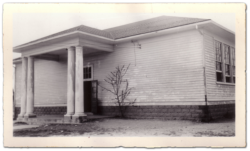 Black and white photograph of the two-room Groveton School. The building has a dark shingled roof with two brick chimneys and a brick foundation. The building is wired for electricity. The wood panel siding is painted white and there is an overhang above the front door held up by four round columns. There is a single door with a transom at the front of the building leading to a central hallway with a classroom on each side. There are large windows along both sides of the building and a single window at the end of the hallway in the rear.