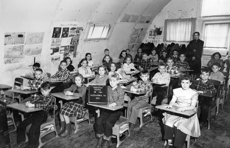 Black and white photograph taken inside one of the Quonset huts at Groveton Elementary School. 29 children and their teacher are pictured. The walls of the building are curved and form a high arched ceiling. The students’ desks have been pushed together in a corner to fit all the children into the photograph. The teacher stands at the back of the room. A boy at one of the desks in front is holding up a sign that reads Groveton School, Grade 3 and 4, Alexandria, Virginia, 1953.