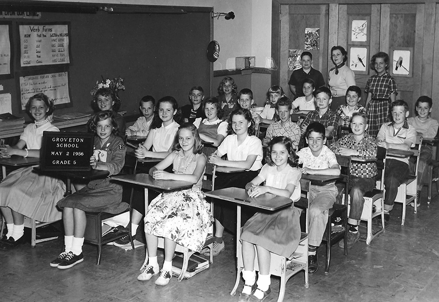 Black and white class photograph showing Mrs. Redd's fifth grade class. All except two of the children are seated at their desks. The desks have been pushed close together to better fit everyone into the picture. Two students are standing at the back of the room with Mrs. Redd. 23 children are pictured. The boys are wearing button down shirts and slacks. The girls are wearing poodle skirts. One girl is wearing a Brownie scout uniform and is holding up a sign that reads Groveton School, May 2, 1956, Grade 5. 