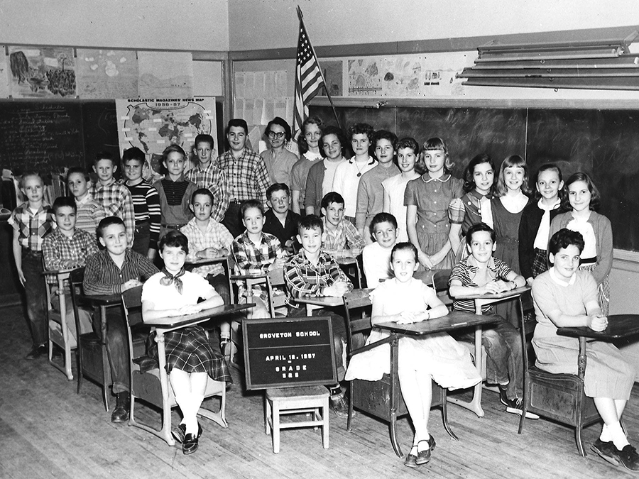 Black and white photograph showing Mrs. Valentine's class comprised of 5th and 6th grade students. 29 children and their teacher are pictured. Approximately half of the children are seated at desks that have been pushed close together. The children who are standing have been arranged along the side and back rows of the desks. Mrs. Valentine is visible in the very back. A sign at the front between two girls reads: Groveton School, April 1957, Grade 5 and 6. The letters on the sign are difficult to read, but the day is either the 12th or the 18th of April.   