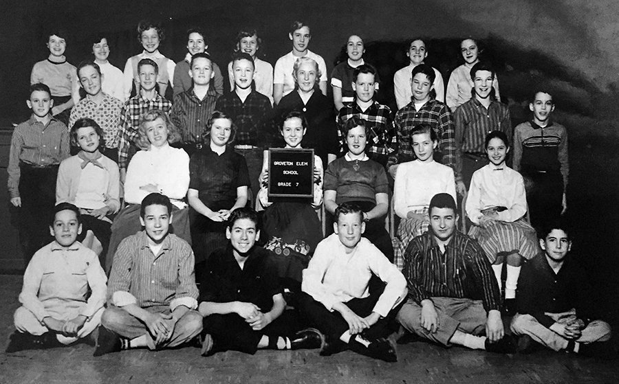 Black and white photograph of Mrs. Hoover's seventh grade class taken during the 1957 to 1958 school year. The children are arranged in four rows with the first row seated on the floor, the second row seated on chairs, the third row standing on the floor, and the fourth row standing on chairs. Mrs. Hoover is standing in the center of the third row. 31 children are pictured, an even mix of boys and girls.
