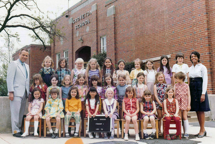 Color photograph from the 1971 to 1972 school year showing Mrs. Brook's Primary grade class of first or second graders. The students pictured are all girls. They are arranged in three rows outside the school near the main entrance. 28 children, Mrs. Brooks, and Principal Zepka are pictured.