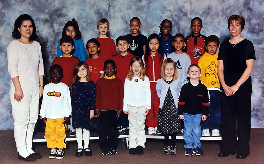 Color photograph showing a kindergarten class portrait. Two teachers and 17 children are shown. The children are arranged in three rows, with the back two rows standing on risers. The teachers stand on opposite sides of the group.