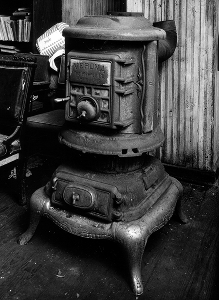 Black and white photograph of a potbelly stove. A potbelly stove is a cast-iron wood-burning stove, round with a bulge in the middle. The name is derived from the resemblance of the stove to that of a fat man's pot belly. They were designed to heat large spaces and were often found in train stations or one-room schoolhouses. The flat top of the fireplace allowed for cooking of food, or the heating of water.