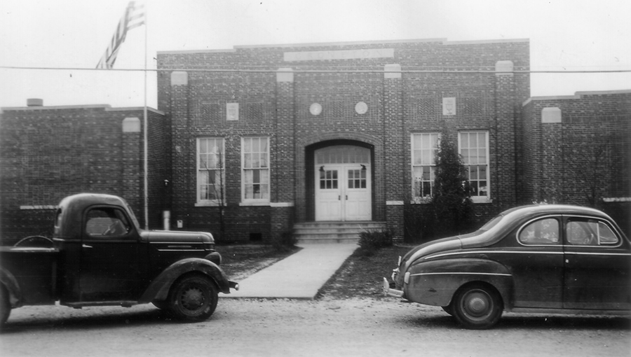 Black and white photograph of Groveton Elementary School, constructed in 1933. The main entrance of the building is shown. The building is a brick structure with two sets of tall narrow windows on both sides of an arched entryway. A concrete sidewalk leads up to the main entrance. Two 1930s-era vehicles, a car and a truck, are parked in front of the building.