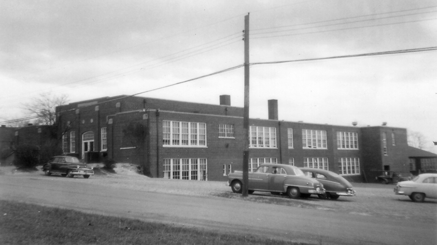 Black and white photograph of the 1933 Groveton Elementary School. The photograph was taken from the opposite side of Memorial Street and shows the front and west side of the building. On this side, the building is two stories tall. It is made of bricks and has several large banks of windows at each classroom. The second addition to the building is complete and the building has more than doubled in size since its original construction. Several cars are parked in a lot next to the school. 