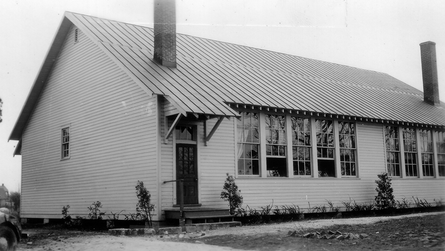 Black and white photograph of the Gum Springs School taken in 1942. The rear of the building is shown. The building has two classrooms and was built using a Rosenwald design, but it was not constructed with Rosenwald Foundation funding. Each classroom is heated with its own stove and there are two brick chimneys protruding from the structure's tin room. Each classroom has a large bank of windows, and its own door to the outside. The sides of the building are covered in white-painted wood siding. The school had no running water or bathrooms. Children used outhouses that are not shown in the photograph. 