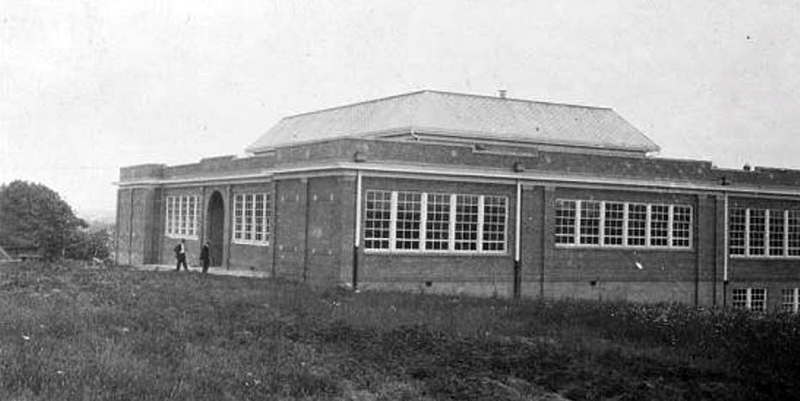 Black and white photograph of Lee-Jackson High School. The school is a two-story brick building built into a hillside so that from the front view of the main entrance the building appears only one-story tall. Large banks of windows line three sides of the building. According to FCPS records, the windows were a frequent target of rock-throwing vandals. There is a raised section at the center of the building with a pitched roof, indicating there may have been an auditorium is this area. The main entrance doors are reached through a tall, brick archway.