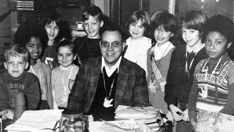 Black and white photograph of Principal Bill Zepka taken in 1981. He is seated at his desk in the library and is surrounded by a large group of smiling students. Nine children are pictured.