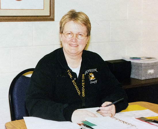 Color photograph of Principal Henderson taken during the 1997 to 1998 school year. She is seated at a table and is looking up from the paperwork in front of her. A pen is in her left hand.