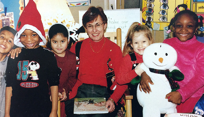 Color photograph of Principal Lamb taken during the 2002 to 2003 school year. She is seated on a brown rocking chair in a classroom with a closed picture book on her lap. She is surrounded by a group of five smiling children, two boys and three girls. Two of the girls, on Lamb's right, are holding a stuffed snowman figure.