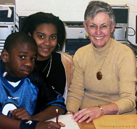 Photograph of Principal Lamb with two students from the 2006 to 2007 Groveton Elementary School yearbook. They are seated at a table with schoolwork in front of them. Two computers and a printer are visible on a table behind them.