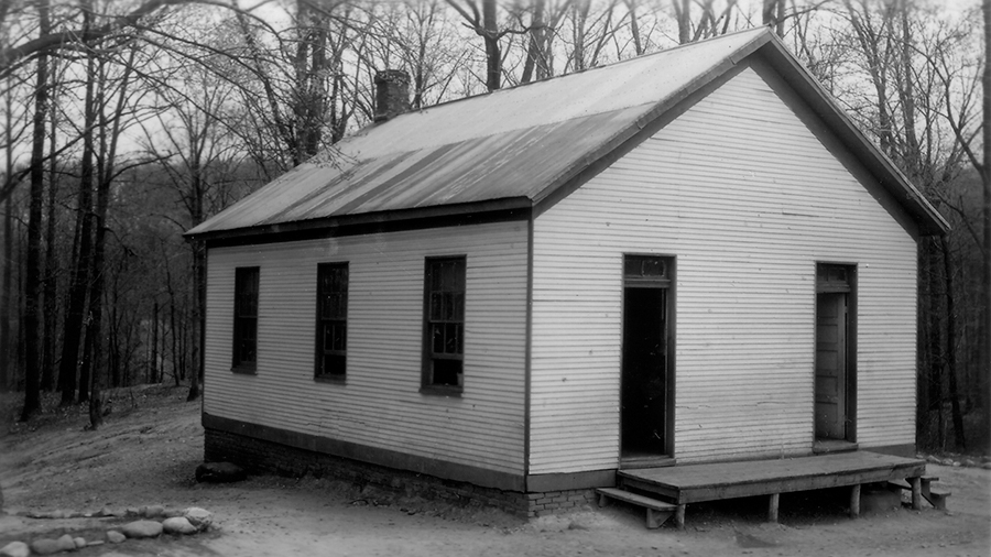 Black and white photograph of the Spring Bank School taken in 1942. It is a one-room schoolhouse with white-painted wood siding on a brick foundation. There are two doors in the front of the building and three windows on each side. A brick chimney can be seen on the rear of the structure. The school is built on a hillside surrounded by trees. The building's roof and siding appear worn with age.