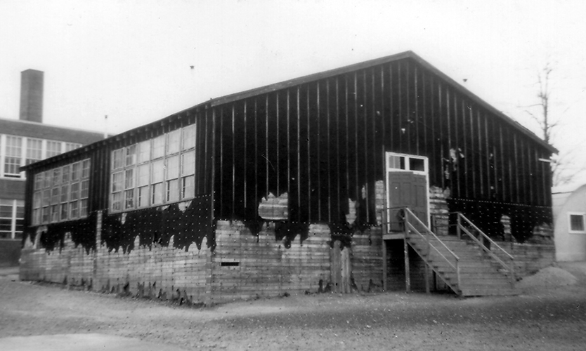 Black and white photograph, taken in 1954, showing the four-classroom temporary building at Groveton. The structure is a wood-frame building with small wood boards used for siding. The siding has been covered with sheets of tarpaper affixed vertically. The tarpaper has been torn off or has worn off toward the bottom of the building. The structure resembles a large shed in shape, and has a central hallway in the middle with two classrooms on either side The building looks very worn and flimsy, and records indicate there were problems with rats requiring the hiring of exterminators. 