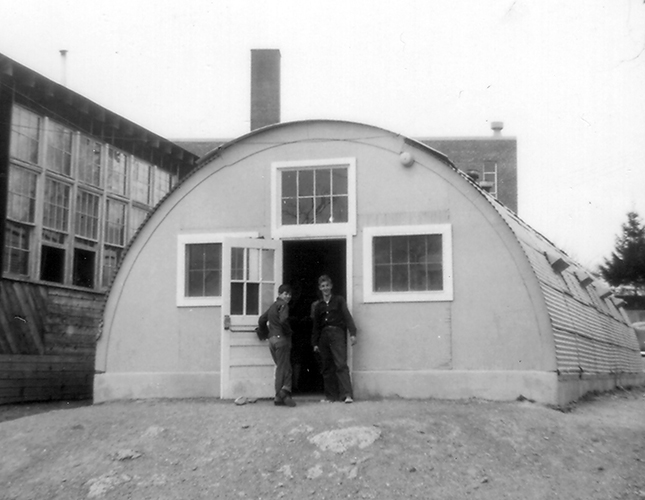 Black and white photograph of one of the Quonset huts used at Groveton Elementary School. The building is made out of metal and is similar in shape to half of a cylinder. The building is approximately 20 feet wide and 48 feet long. The sides are covered with corrugated steel sheets. The building has three windows and a door at one end, and four windows on each of the long sides. Two students are pictured standing in the open doorway to the Quonset hut. The tarpaper building is to the left of the hut and the brick school building is visible behind it.